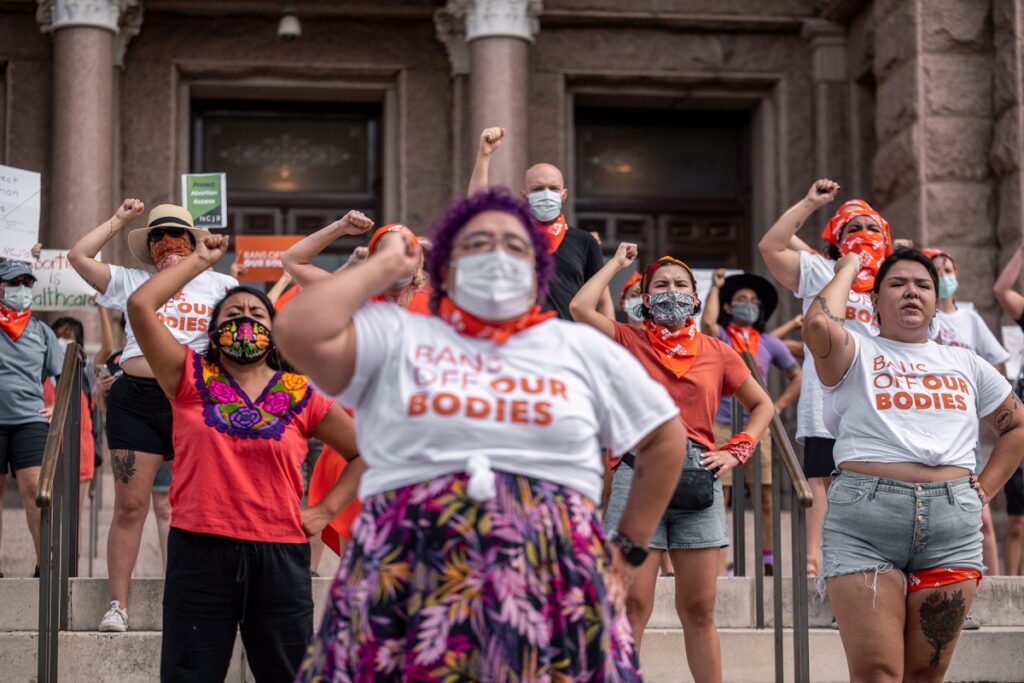 Pro-choice protesters stand in front of the Texas State capitol with their right fist up in the air, and some of them were a white t-shirt with orange text that says, "Bans Off Our Bodies."